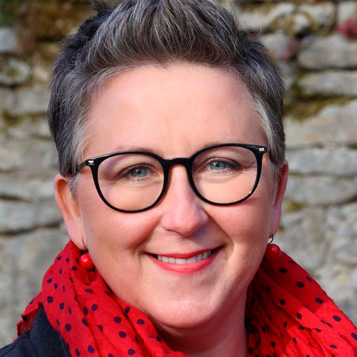 Eine Frau mittleren Alters mit kurzem Haar, schwarzer Brille und rotem Schal mit schwarzen Punkten. Sie lächelt freundlich.