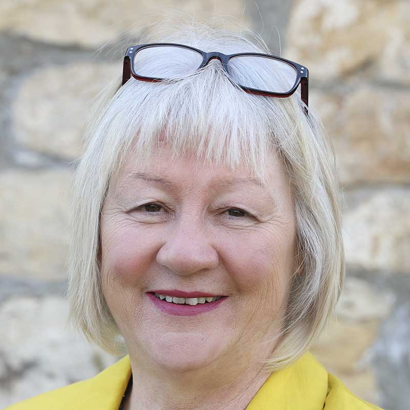 Eine Frau mit kurzen, blonden Haaren und schwarzen Gläsern auf dem Kopf lächelt in die Kamera. Sie trägt einen gelben Pullover.