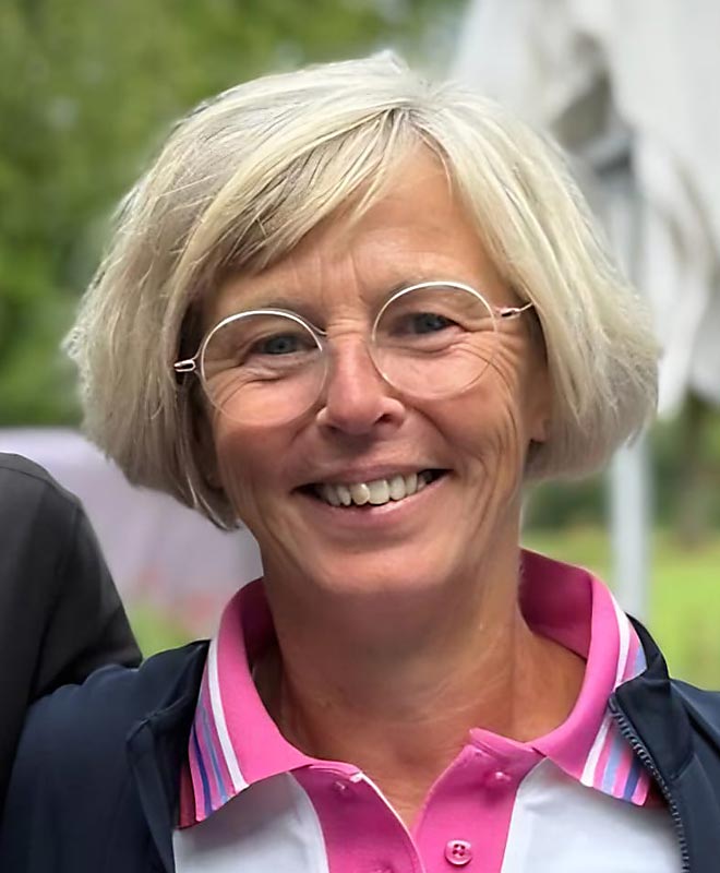 Eine lächelnde Frau mit kurzen, blonden Haaren und Brille. Sie trägt ein weißes Hemd mit rosa Kragen.