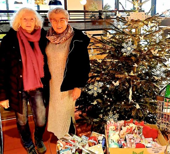 Zwei Frauen stehen neben einem Weihnachtsbaum und Spendendosen. Sie lächeln in die Kamera.