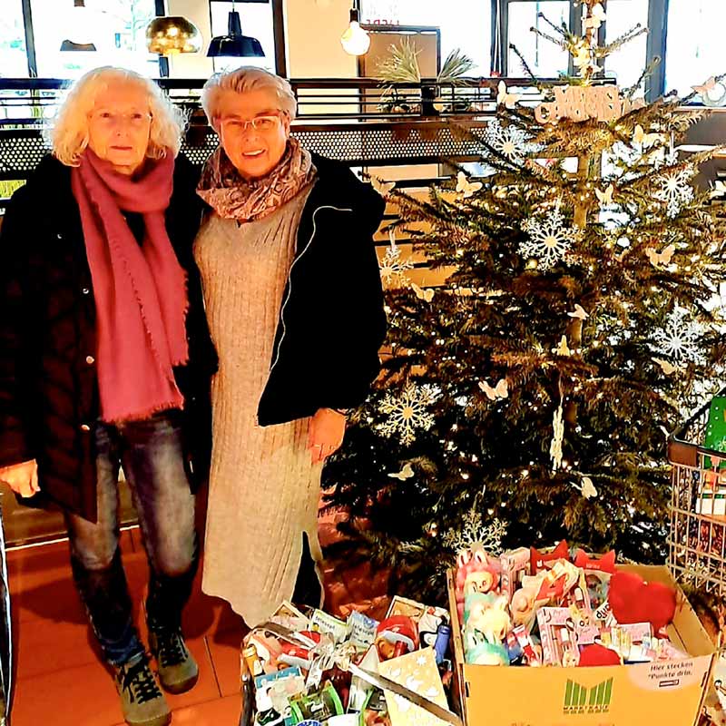 Zwei Frauen stehen neben einem Weihnachtsbaum und Spendendosen. Sie lächeln in die Kamera.