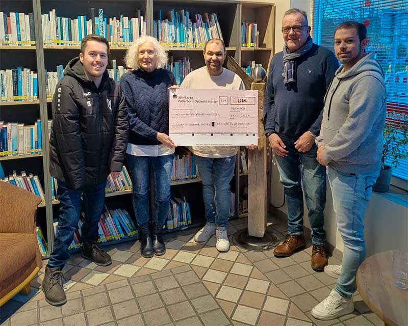 Eine Gruppe von fünf Personen hält einen großen Scheck in einem Raum mit Büchern im Hintergrund. Der Scheck scheint eine Spende oder eine Auszeichnung zu sein.