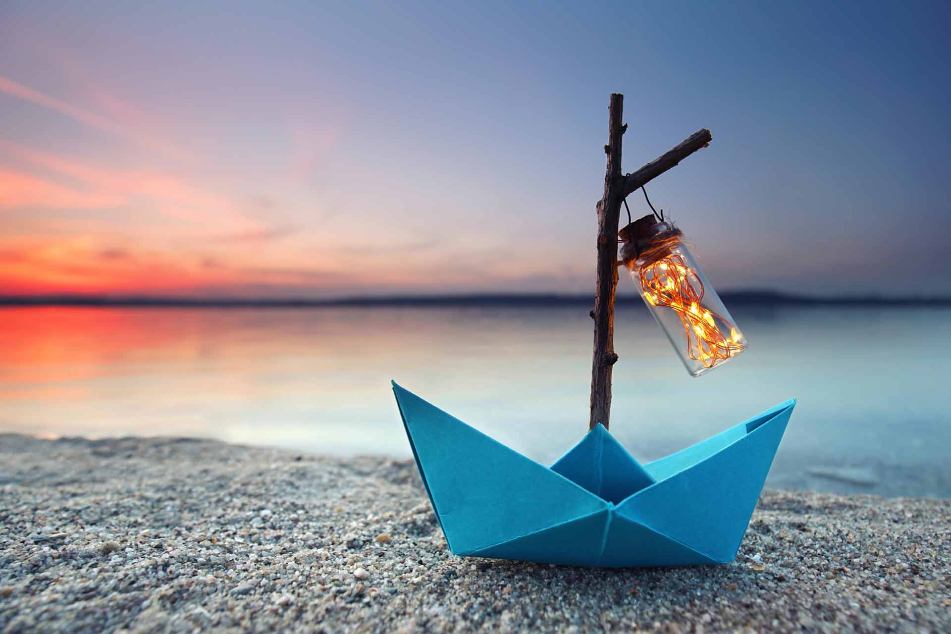 Ein blau gefaltetes Papierboot mit einer leuchtenden Laterne auf einem Sandstrand bei Sonnenuntergang.